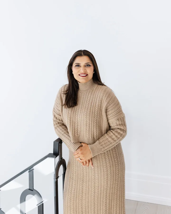 Ravi Gill in a camel knit dress beside a modern staircase in a bright white home interior